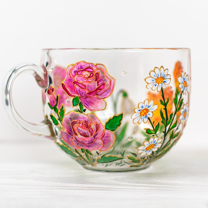K&ouml;nnte beinhalten: Eine transparente Teetasse mit Henkel, handbemalt mit rosa Rosen, wei&szlig;en G&auml;nsebl&uuml;mchen und orangefarbenen Blumen. Das florale Design ist goldfarben umrandet, mit gr&uuml;nen Bl&auml;ttern. Die Tasse steht auf einer wei&szlig;en Oberfl&auml;che.