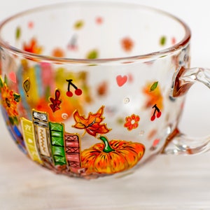 Puede incluir: Una taza de cristal transparente con dise&ntilde;os pintados a mano de una calabaza naranja brillante, una pila de libros coloridos, hojas de oto&ntilde;o, peque&ntilde;as cerezas rojas y diminutos corazones rosas. La taza tiene un asa curva.