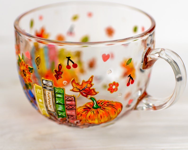 Puede incluir: Una taza de cristal transparente con dise&ntilde;os pintados a mano de una calabaza naranja brillante, una pila de libros coloridos, hojas de oto&ntilde;o, peque&ntilde;as cerezas rojas y diminutos corazones rosas. La taza tiene un asa curva.
