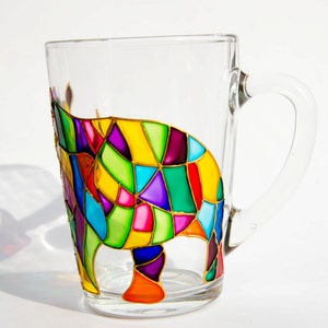 May include: A clear glass mug with a colorful, geometric design of an animal. The design features a patchwork of vibrant colors including yellow, green, purple, red, and blue, outlined in gold. The mug has a handle.