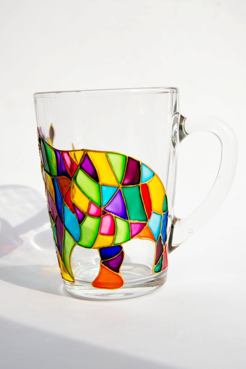 May include: A clear glass mug with a colorful, geometric design of an animal. The design features a patchwork of vibrant colors including yellow, green, purple, red, and blue, outlined in gold. The mug has a handle.