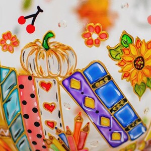 Puede incluir: Una superficie de cristal decorada con coloridas ilustraciones pintadas que incluyen una calabaza blanca, un girasol amarillo, cerezas rojas y varios libros en tonos azules, morados y rosas con lunares y corazones, junto a l&aacute;pices de colores.