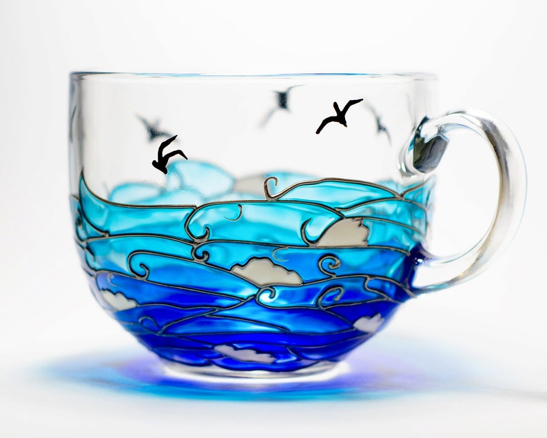 May include: A clear glass teacup with a painted ocean wave design in shades of blue and white. Black silhouettes of seagulls are flying above the waves. The cup has a clear handle and sits on a white surface.