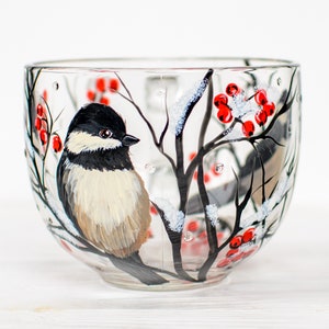 May include: A clear glass mug featuring a hand-painted design of a black-capped chickadee perched on a dark branch. The scene includes clusters of bright red berries and white snow accents on the branches, creating a winter-themed illustration.
