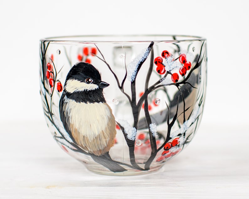 May include: A clear glass mug featuring a hand-painted design of a black-capped chickadee perched on a dark branch. The scene includes clusters of bright red berries and white snow accents on the branches, creating a winter-themed illustration.