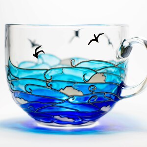 Peut inclure: Une tasse en verre transparent orn&eacute;e d'un motif peint &agrave; la main repr&eacute;sentant des vagues oc&eacute;aniques tourbillonnantes dans diverses nuances de bleu, avec des d&eacute;tails d'&eacute;cume blanche et des silhouettes noires d'oiseaux volant au-dessus.