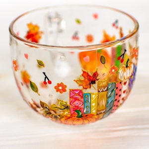 Puede incluir: Una taza de cristal transparente decorada con motivos pintados a mano que incluyen hojas de oto&ntilde;o coloridas, calabazas naranjas, peque&ntilde;as cerezas rojas, flores rosas y una pila de libros vibrantes con detalles en tonos naranja, rojo y azul.