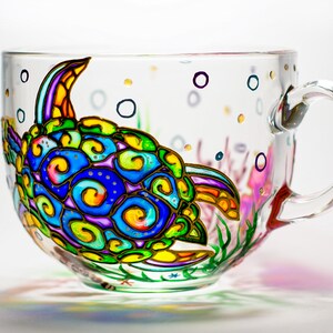 May include: A clear glass teacup with a colorful, hand-painted sea turtle design. The turtle features blue, green, yellow, and purple swirls. The cup also has painted bubbles and aquatic plants. The handle is clear glass.