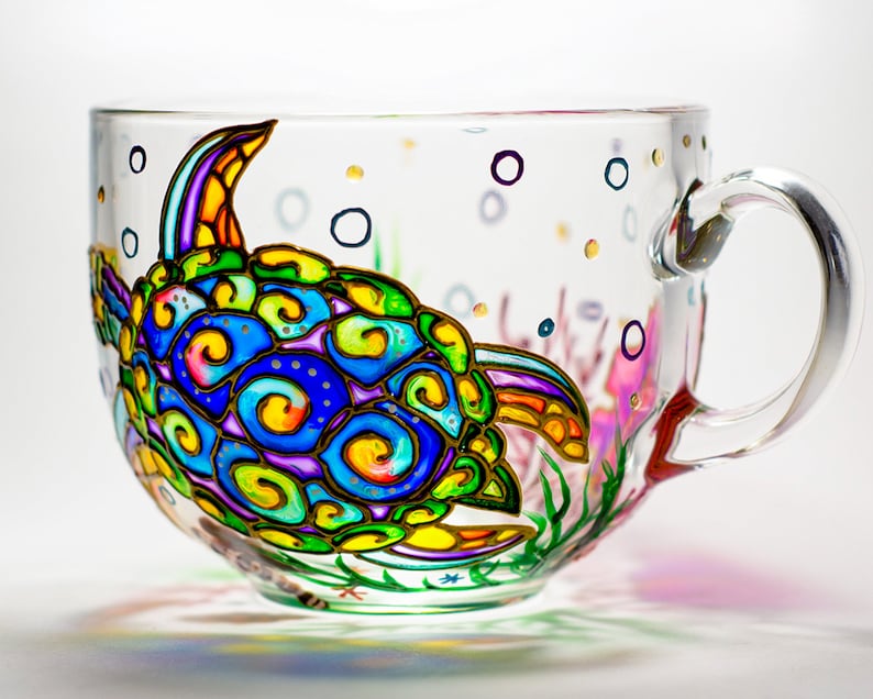 May include: A clear glass teacup with a colorful, hand-painted sea turtle design. The turtle features blue, green, yellow, and purple swirls. The cup also has painted bubbles and aquatic plants. The handle is clear glass.
