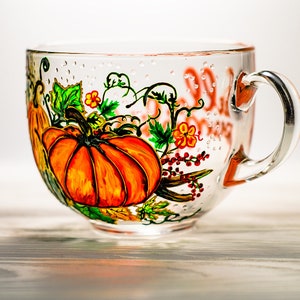 May include: A clear glass mug featuring a hand-painted design of bright orange pumpkins, green leaves, and small red berries. The glass surface is decorated with subtle raised dots, and the handle is attached to the right side of the mug.