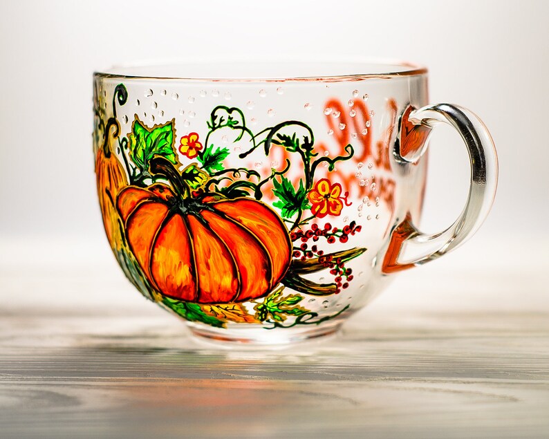 May include: A clear glass mug featuring a hand-painted design of bright orange pumpkins, green leaves, and small red berries. The glass surface is decorated with subtle raised dots, and the handle is attached to the right side of the mug.