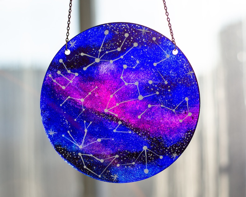 May include: A round glass bauble with a blue and purple galaxy design and white constellations painted on it. The bauble is hanging from a silver chain.