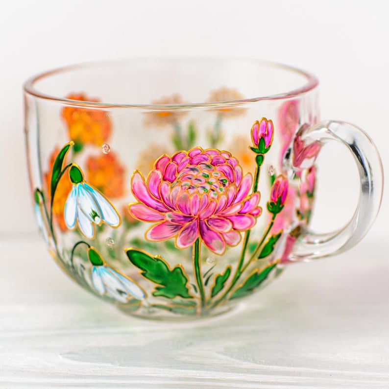K&ouml;nnte beinhalten: Eine transparente Teetasse mit einem gro&szlig;en rosa-goldenen Blumendesign. Die Tasse hat einen Henkel und ist mit verschiedenen Blumen in Rosa-, Orange- und Wei&szlig;t&ouml;nen mit gr&uuml;nen Bl&auml;ttern verziert.