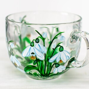 May include: A clear glass mug featuring a hand-painted design of white snowdrop flowers with green stems and leaves. The floral artwork is detailed with blue accents on the petals and yellow centers, wrapping around the exterior of the transparent cup.