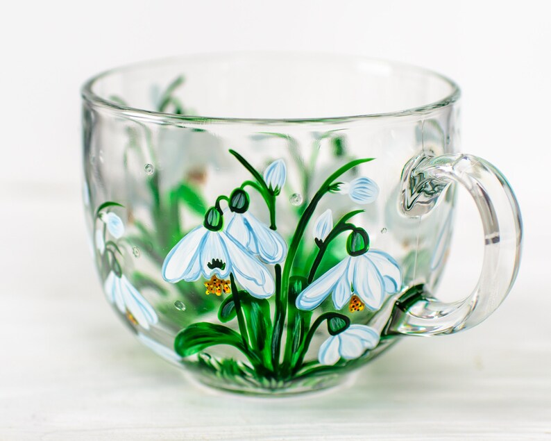 May include: A clear glass mug featuring a hand-painted design of white snowdrop flowers with green stems and leaves. The floral artwork is detailed with blue accents on the petals and yellow centers, wrapping around the exterior of the transparent cup.