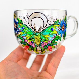 May include: A clear glass teacup with a handle, hand-painted with a colorful butterfly design. The butterfly has green, yellow, and blue wings, surrounded by blue flowers and pink berries. A crescent moon and stars are also painted on the cup.