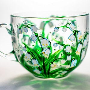 Puede incluir: Una taza de t&eacute; de vidrio transparente con asa, adornada con flores blancas y azules en forma de campana pintadas a mano y hojas verdes. El dise&ntilde;o floral envuelve la taza, creando un aspecto delicado. Peque&ntilde;as gotas transparentes pintadas sobre el vidrio.