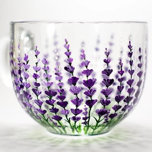May include: A clear glass mug featuring a hand-painted design of purple lavender sprigs with green stems. The floral pattern wraps around the exterior of the cup, showcasing various shades of violet and deep purple petals against the transparent glass.
