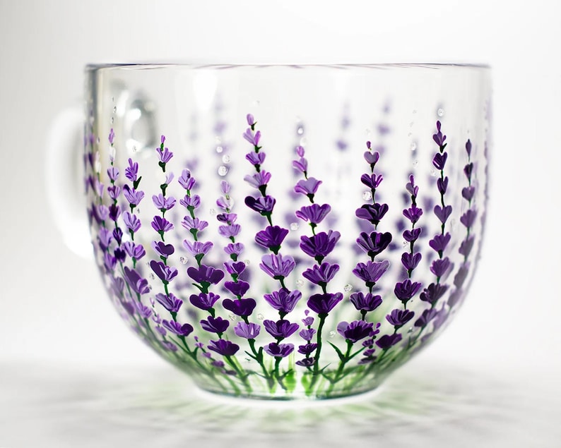 May include: A clear glass mug featuring a hand-painted design of purple lavender sprigs with green stems. The floral pattern wraps around the exterior of the cup, showcasing various shades of violet and deep purple petals against the transparent glass.