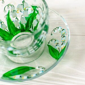 May include: A clear glass teacup and saucer set, hand-painted with a lily of the valley design. The design features white bell-shaped flowers with yellow centers and green leaves. The teacup and saucer are on a white wooden surface.