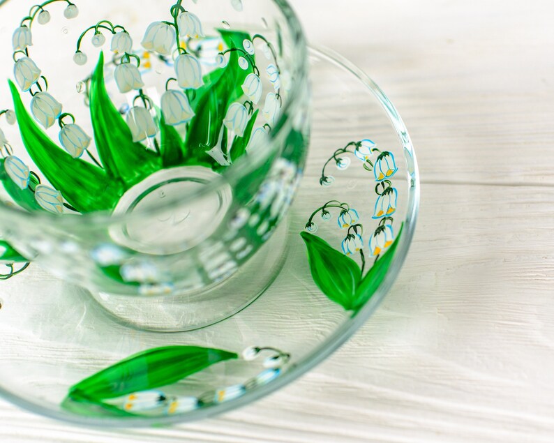May include: A clear glass teacup and saucer set, hand-painted with a lily of the valley design. The design features white bell-shaped flowers with yellow centers and green leaves. The teacup and saucer are on a white wooden surface.