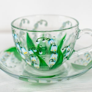 May include: A clear glass teacup and saucer set. The cup and saucer are decorated with a hand-painted design of white and yellow lily of the valley flowers with green leaves. The cup has a handle and sits on a matching saucer.