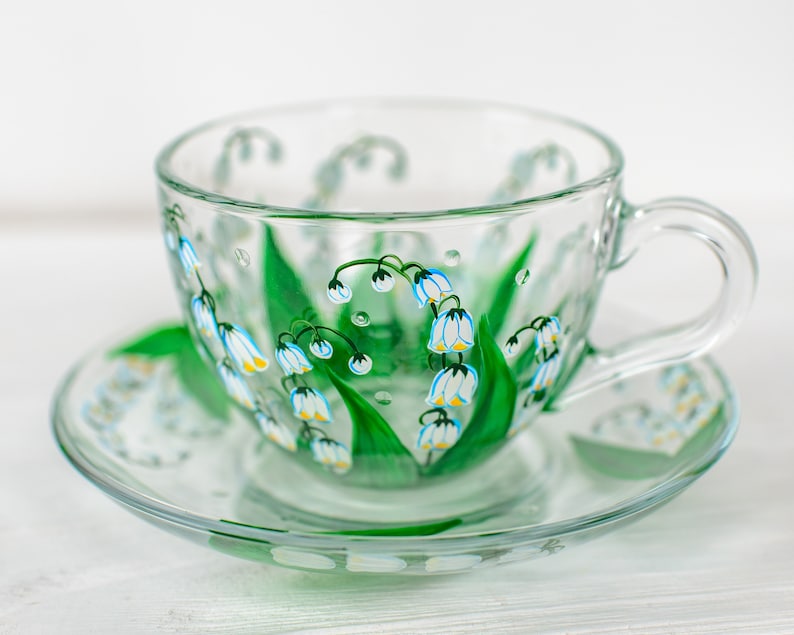May include: A clear glass teacup and saucer set. The cup and saucer are decorated with a hand-painted design of white and yellow lily of the valley flowers with green leaves. The cup has a handle and sits on a matching saucer.