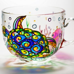 May include: A clear glass teacup with a hand-painted sea turtle design. The turtle features vibrant colors including blue, green, yellow, and purple. The cup also has painted bubbles and coral accents, creating an underwater theme.