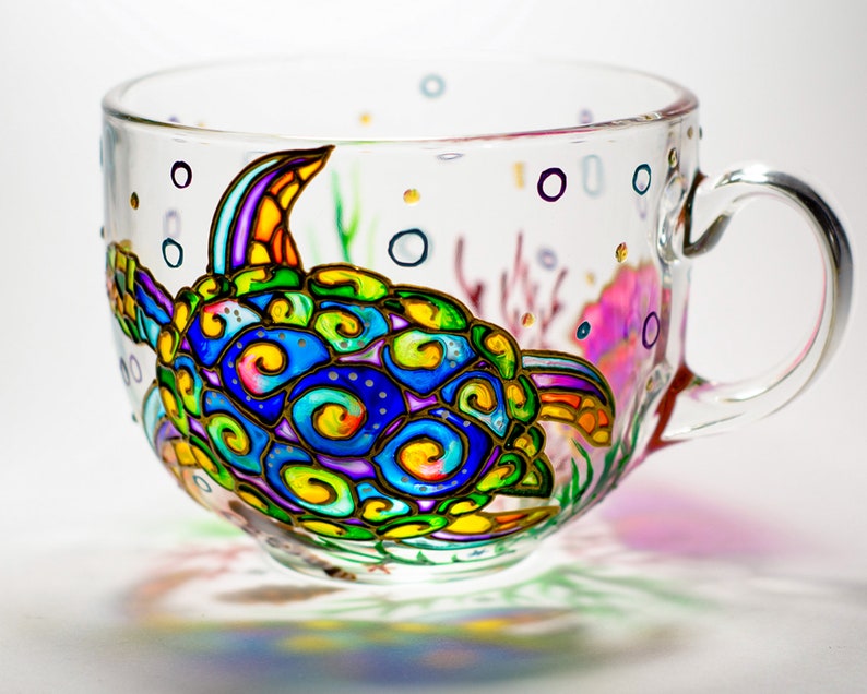 May include: A clear glass teacup with a hand-painted sea turtle design. The turtle features vibrant colors including blue, green, yellow, and purple. The cup also has painted bubbles and coral accents, creating an underwater theme.