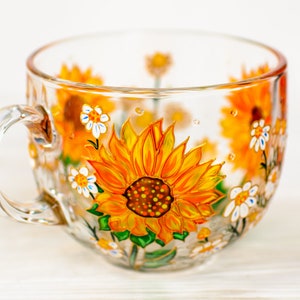 Op de afbeelding: Een doorzichtige glazen mok met handgeschilderde gele en oranje zonnebloemen, groene bladeren en kleine witte bloemetjes. Het ontwerp loopt rond de buitenkant van de beker, die aan de linkerkant is voorzien van een gebogen handvat.