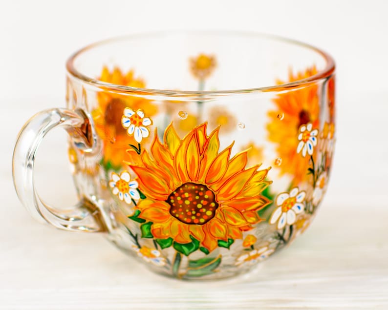 Op de afbeelding: Een doorzichtige glazen mok met handgeschilderde gele en oranje zonnebloemen, groene bladeren en kleine witte bloemetjes. Het ontwerp loopt rond de buitenkant van de beker, die aan de linkerkant is voorzien van een gebogen handvat.