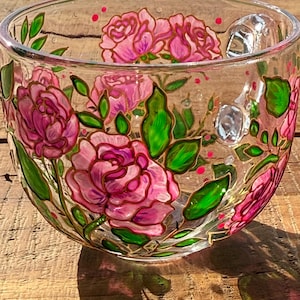 Puede incluir: Un cuenco de vidrio transparente con rosas rosas pintadas a mano, hojas verdes y detalles dorados. El dise&ntilde;o floral rodea el cuenco, con peque&ntilde;os puntos rojos. El cuenco est&aacute; sobre una superficie de madera.