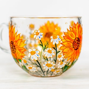 Op de afbeelding: Een doorzichtige glazen mok met een handgeschilderd ontwerp van levendige gele zonnebloemen en kleine witte madeliefjes met gele harten. De bloemenprint loopt rondom de buitenkant van de beker en toont groene stengels en bladeren op het glas.