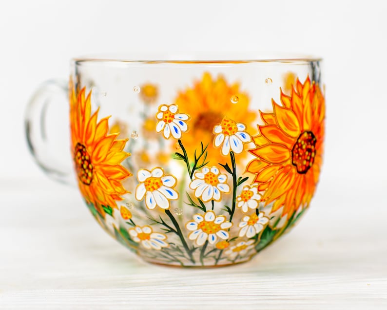 Op de afbeelding: Een doorzichtige glazen mok met een handgeschilderd ontwerp van levendige gele zonnebloemen en kleine witte madeliefjes met gele harten. De bloemenprint loopt rondom de buitenkant van de beker en toont groene stengels en bladeren op het glas.