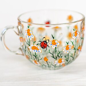 Puede incluir: Una taza de t&eacute; de vidrio transparente con asa, pintada a mano con un dise&ntilde;o floral de margaritas blancas, centros naranjas y tallos verdes. Una mariquita roja tambi&eacute;n est&aacute; pintada en la taza. La taza est&aacute; sobre una superficie blanca.