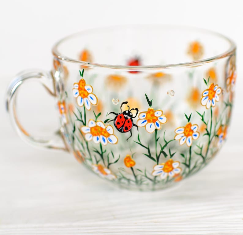 Puede incluir: Una taza de t&eacute; de vidrio transparente con asa, pintada a mano con un dise&ntilde;o floral de margaritas blancas, centros naranjas y tallos verdes. Una mariquita roja tambi&eacute;n est&aacute; pintada en la taza. La taza est&aacute; sobre una superficie blanca.