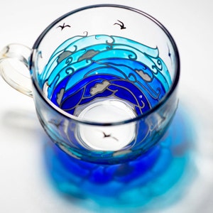 Puede incluir: Una taza de t&eacute; de vidrio transparente con un dise&ntilde;o de olas oce&aacute;nicas pintadas en tonos azules. El dise&ntilde;o incluye olas estilizadas, nubes y peque&ntilde;as siluetas de p&aacute;jaros negros. La taza tiene un asa y proyecta una sombra azul.