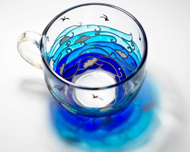 Puede incluir: Una taza de t&eacute; de vidrio transparente con un dise&ntilde;o de olas oce&aacute;nicas pintadas en tonos azules. El dise&ntilde;o incluye olas estilizadas, nubes y peque&ntilde;as siluetas de p&aacute;jaros negros. La taza tiene un asa y proyecta una sombra azul.