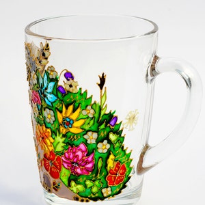 May include: A clear glass mug with a colorful floral design painted on the side. The flowers are painted in shades of red, pink, yellow, blue, green, and white. The mug has a gold rim and handle.