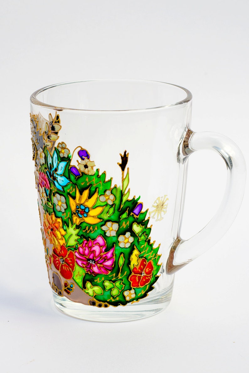 May include: A clear glass mug with a colorful floral design painted on the side. The flowers are painted in shades of red, pink, yellow, blue, green, and white. The mug has a gold rim and handle.