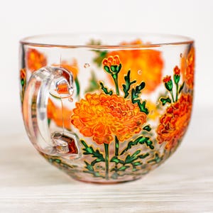 May include: A clear glass teacup with a handle, adorned with hand-painted orange marigold flowers and green leaves. The floral design is outlined in gold, adding a touch of elegance to the cup's exterior. The cup is ideal for tea or coffee.