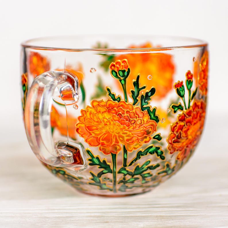 May include: A clear glass teacup with a handle, adorned with hand-painted orange marigold flowers and green leaves. The floral design is outlined in gold, adding a touch of elegance to the cup's exterior. The cup is ideal for tea or coffee.