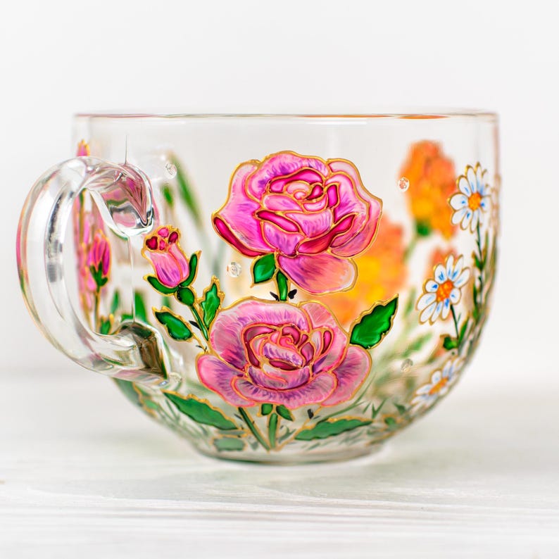 K&ouml;nnte beinhalten: Transparente Teetasse mit gro&szlig;em Henkel, handbemalt mit rosa Rosen, gr&uuml;nen Bl&auml;ttern und goldenen Akzenten. Die Tasse zeigt auch orangefarbene und wei&szlig;e, g&auml;nsebl&uuml;mchenartige Blumen. Das Design befindet sich auf der Au&szlig;enseite der Tasse.