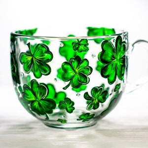 May include: A clear glass mug with a large handle, decorated with hand-painted green shamrocks. The shamrocks vary in size and are evenly spaced around the mug. The mug is ideal for serving beverages.
