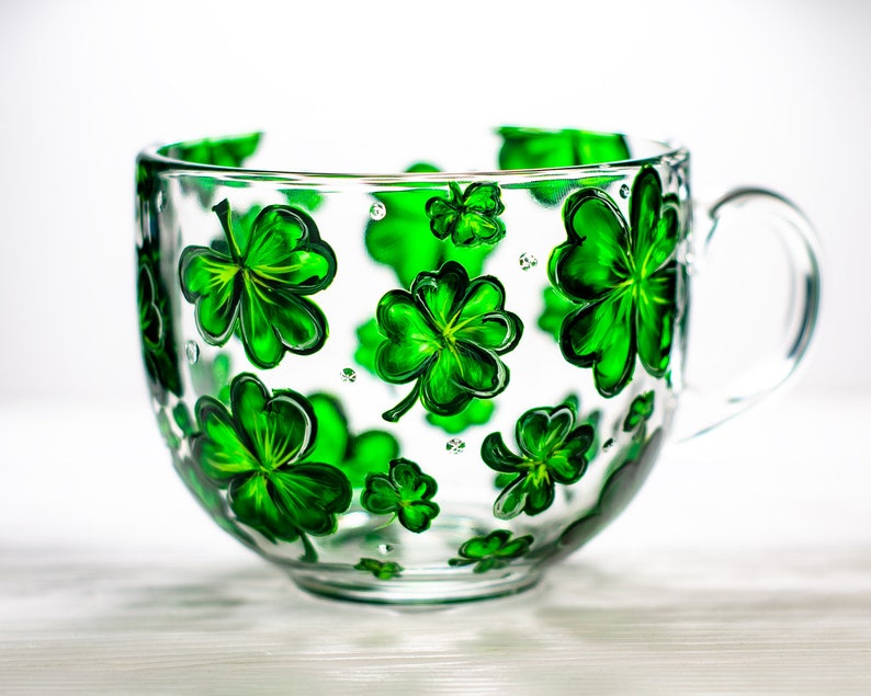 May include: A clear glass mug with a large handle, decorated with hand-painted green shamrocks. The shamrocks vary in size and are evenly spaced around the mug. The mug is ideal for serving beverages.