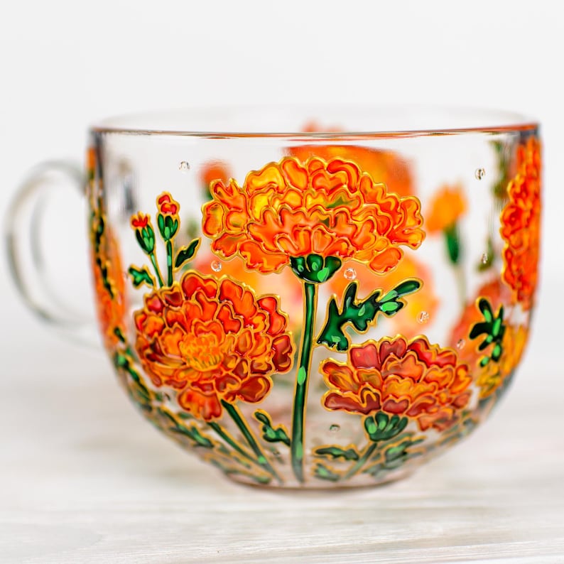 May include: A clear glass teacup with a handle, adorned with hand-painted orange and yellow marigold flowers. The floral design is outlined in gold, with green stems and leaves. The cup is set against a white background.