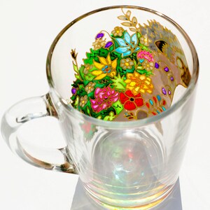 May include: A clear glass mug with a colorful floral design painted on the side. The design features a variety of flowers in shades of pink, yellow, blue, green, and red. The flowers are surrounded by gold accents.