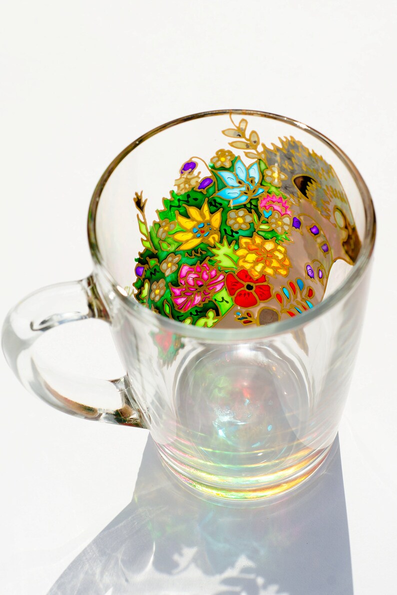 May include: A clear glass mug with a colorful floral design painted on the side. The design features a variety of flowers in shades of pink, yellow, blue, green, and red. The flowers are surrounded by gold accents.