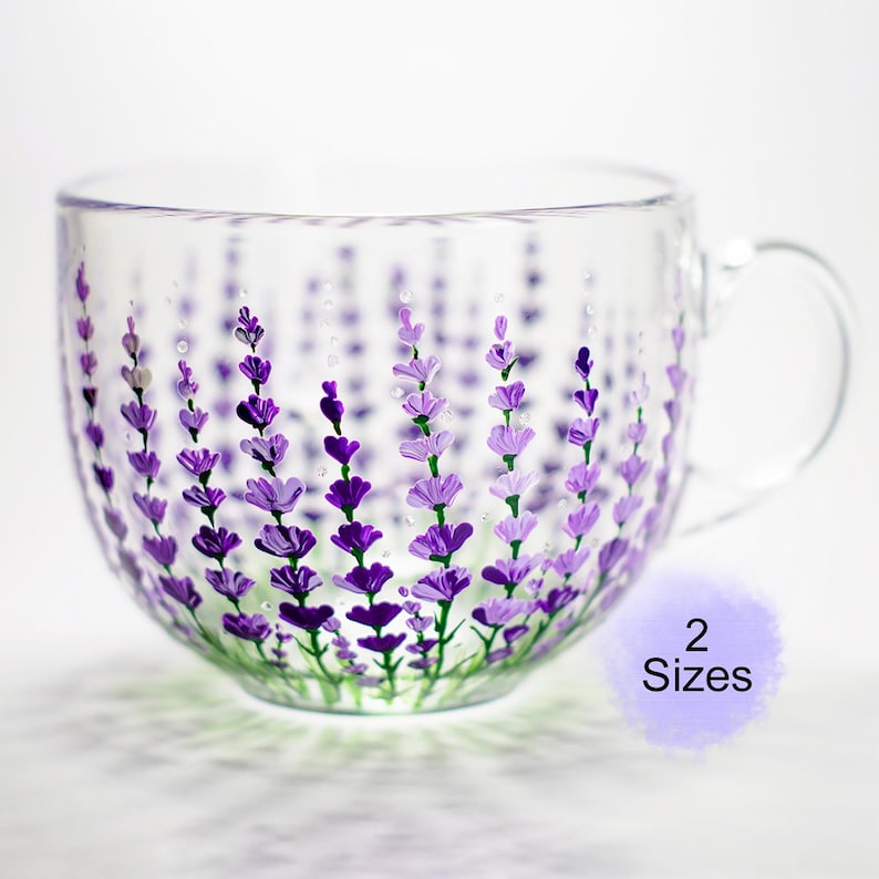 May include: A clear glass teacup with a handle, hand-painted with purple lavender sprigs. The lavender design is painted on the outside of the cup, with green stems at the base. The cup is available in 2 sizes.