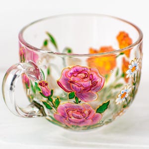K&ouml;nnte beinhalten: Eine transparente Teetasse mit Henkel, handbemalt mit rosa Rosen, orangefarbenen Blumen und wei&szlig;en G&auml;nsebl&uuml;mchen. Das florale Design ist goldfarben umrandet, mit gr&uuml;nen Bl&auml;ttern und Stielen. Die Tasse steht auf einer wei&szlig;en Oberfl&auml;che.